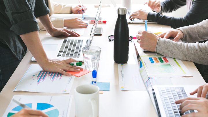 Hands around a meeting table covered with printed charts and laptops